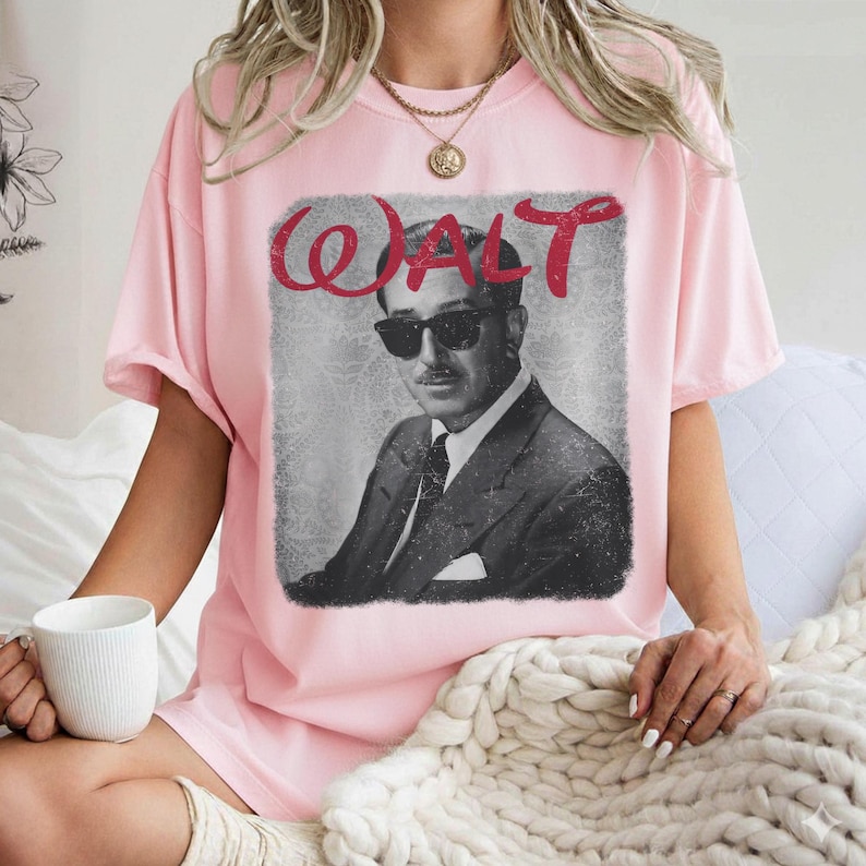 May include: Pink t-shirt featuring a black and white image of a man in a suit and sunglasses, with the name "WALT" in red script above the image. The shirt has short sleeves and a crew neck.
