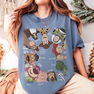 K&ouml;nnte beinhalten: Hellblaues T-Shirt mit einem farbenfrohen Aufdruck verschiedener Charaktere aus dem Animationsfilm Toy Story. Das Design zeigt Woody, Buzz Lightyear und andere Charaktere mit ihren Namen darunter.