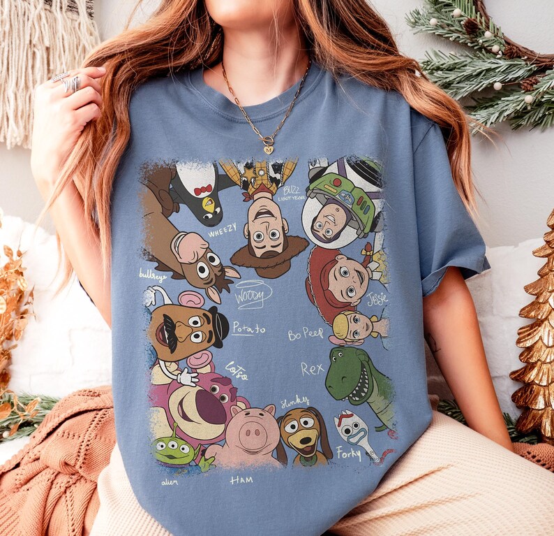 K&ouml;nnte beinhalten: Hellblaues T-Shirt mit einem farbenfrohen Aufdruck verschiedener Charaktere aus dem Animationsfilm Toy Story. Das Design zeigt Woody, Buzz Lightyear und andere Charaktere mit ihren Namen darunter.