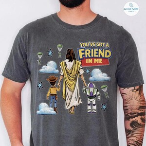 May include: Dark gray t-shirt featuring a graphic design with the text "YOU'VE GOT A FRIEND IN ME". The design includes illustrations of Jesus, Woody, and Buzz Lightyear, along with clouds, stars, and parachutes.