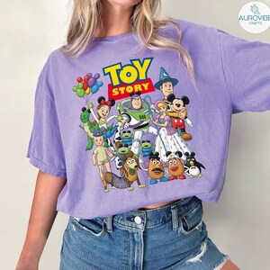 Op de afbeelding: Een paars T-shirt met een kleurrijke afbeelding van personages uit de animatiefilm "Toy Story". Het ontwerp bevat Buzz Lightyear, Woody, Mickey Mouse en andere personages. De tekst "TOY STORY" wordt in geel en rood weergegeven.
