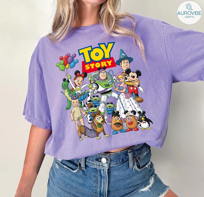 Op de afbeelding: Een paars T-shirt met een kleurrijke afbeelding van personages uit de animatiefilm "Toy Story". Het ontwerp bevat Buzz Lightyear, Woody, Mickey Mouse en andere personages. De tekst "TOY STORY" wordt in geel en rood weergegeven.