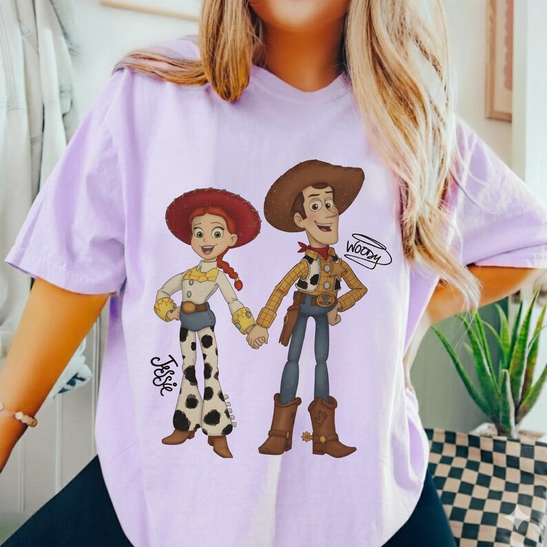 Może przedstawiać: Jasnofioletowa koszulka z kresk&oacute;wkową ilustracją Jessie i Woody'ego z Toy Story, trzymających się za ręce. Jessie ma na sobie czerwony kapelusz, białą koszulę i spodnie w krowie łaty. Woody ma na sobie kapelusz kowbojski, kamizelkę i buty. Na koszulce widnieje napis "Woody".