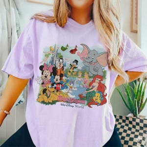 Puede incluir: Una camiseta de color lila claro con un gr&aacute;fico colorido de varios personajes de Disney, incluyendo Mickey Mouse, Minnie Mouse, Donald Duck y Ariel. El gr&aacute;fico tambi&eacute;n incluye el texto "Magic Kingdom Walt Disney World".