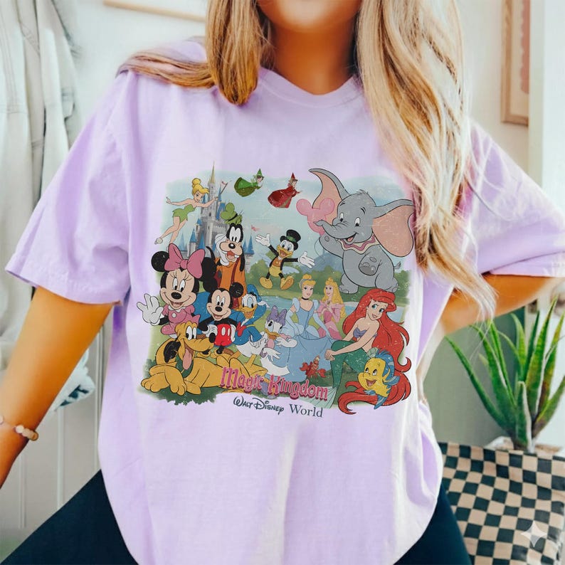 Puede incluir: Una camiseta de color lila claro con un gr&aacute;fico colorido de varios personajes de Disney, incluyendo Mickey Mouse, Minnie Mouse, Donald Duck y Ariel. El gr&aacute;fico tambi&eacute;n incluye el texto "Magic Kingdom Walt Disney World".