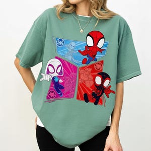 May include: Sage green t-shirt featuring a colorful graphic of Spider-Man characters. The design includes Spider-Man, Spider-Gwen, and other characters in a geometric pattern. The shirt has short sleeves and a crew neck.