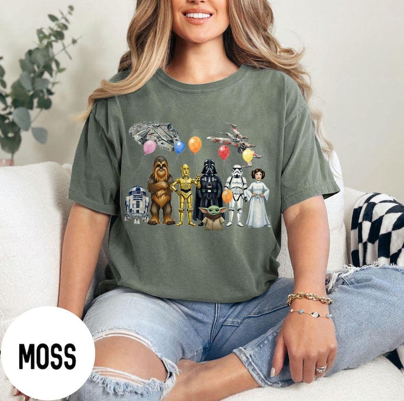 Peut inclure: T-shirt vert mousse avec un motif Star Wars. Le design comprend des personnages, des vaisseaux spatiaux et des ballons. Le mot "MOSS" est imprim&eacute; dans un cercle blanc en bas &agrave; gauche.
