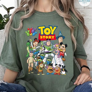 Op de afbeelding: Olijfgroen T-shirt met een kleurrijke afbeelding van Toy Story-personages, waaronder Woody, Buzz Lightyear en Mickey Mouse. De woorden "TOY STORY" staan in een blauw en rood lettertype. Een leuk, speels ontwerp.