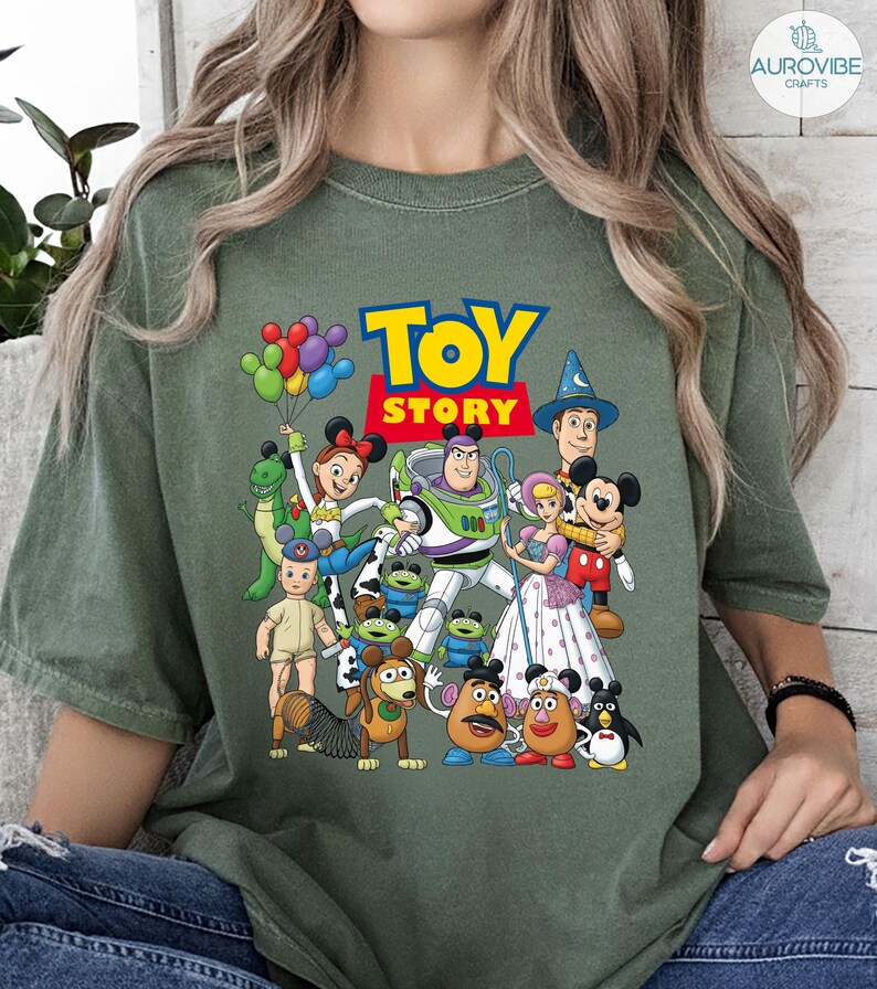Op de afbeelding: Olijfgroen T-shirt met een kleurrijke afbeelding van Toy Story-personages, waaronder Woody, Buzz Lightyear en Mickey Mouse. De woorden "TOY STORY" staan in een blauw en rood lettertype. Een leuk, speels ontwerp.