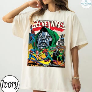 May include: Ivory-colored t-shirt featuring a comic book graphic with the text "SECRET WARS." The graphic depicts various superheroes and villains in action, including Spider-Man, Captain America, and Doctor Doom. The design is in a vintage comic book style.