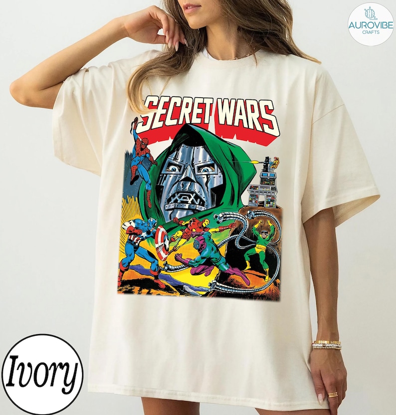 May include: Ivory-colored t-shirt featuring a comic book graphic with the text "SECRET WARS." The graphic depicts various superheroes and villains in action, including Spider-Man, Captain America, and Doctor Doom. The design is in a vintage comic book style.
