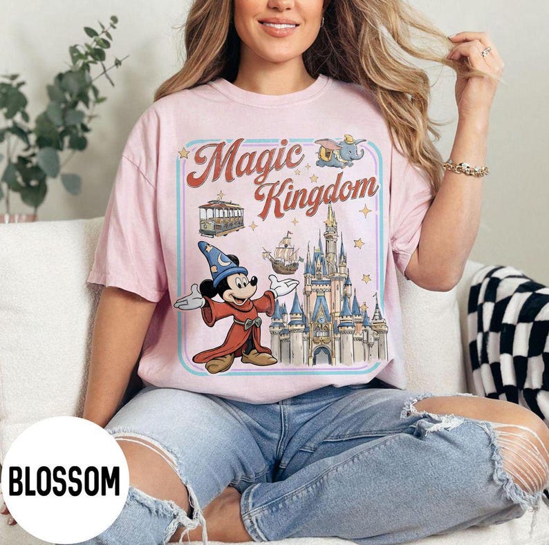 May include: Pink t-shirt with a graphic of Mickey Mouse in a sorcerer's outfit, the words "Magic Kingdom", a castle, and other illustrations. The shirt is a light pink color.