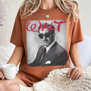 May include: A rust-colored t-shirt featuring a black and white image of a man in sunglasses and a suit. The word "WALT" is written in red cursive above the image. The shirt is a relaxed fit.
