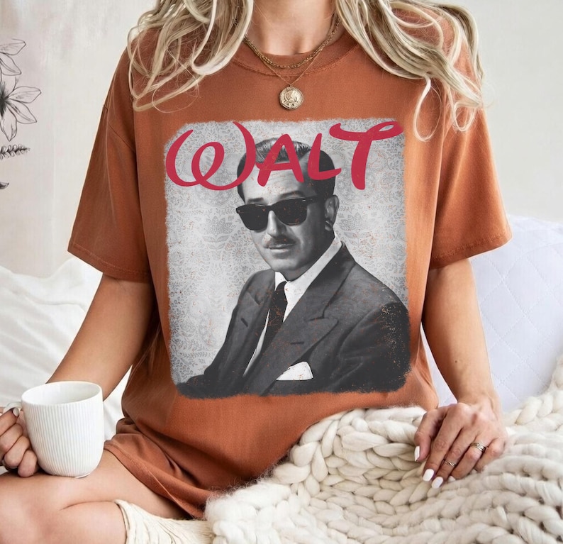 May include: A rust-colored t-shirt featuring a black and white image of a man in sunglasses and a suit. The word "WALT" is written in red cursive above the image. The shirt is a relaxed fit.