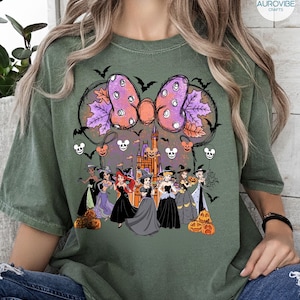 Może przedstawiać: Zielona koszulka z motywem Halloween. Grafika zawiera dużą kokardę z fioletowymi i pomarańczowymi akcentami, nietoperze, dynie i księżniczki Disneya w kostiumach czarownic. W tle znajduje się zamek.