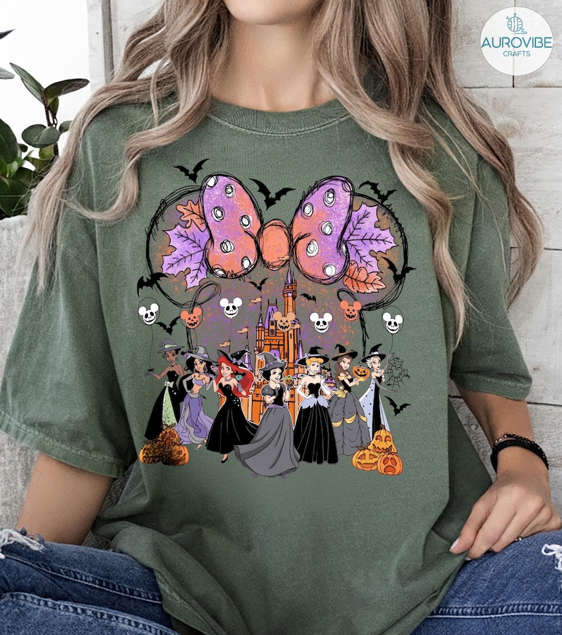 Może przedstawiać: Zielona koszulka z motywem Halloween. Grafika zawiera dużą kokardę z fioletowymi i pomarańczowymi akcentami, nietoperze, dynie i księżniczki Disneya w kostiumach czarownic. W tle znajduje się zamek.
