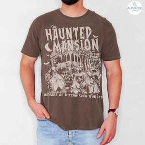 May include: Brown t-shirt featuring a graphic of a haunted mansion with the text "The Haunted Mansion" and "Beware of Hitchhiking Ghosts." The design includes a spooky house, ghosts, and a crescent moon.