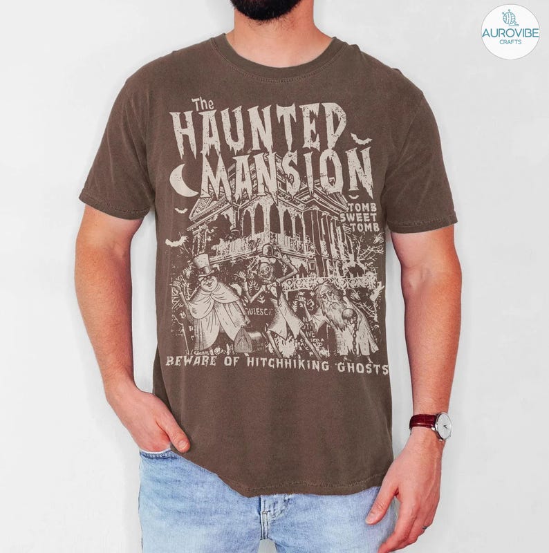 May include: Brown t-shirt featuring a graphic of a haunted mansion with the text "The Haunted Mansion" and "Beware of Hitchhiking Ghosts." The design includes a spooky house, ghosts, and a crescent moon.