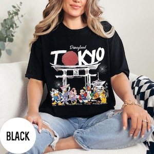 Puede incluir: Camiseta negra con un dise&ntilde;o de Disneyland Tokio. El dise&ntilde;o incluye la palabra "TOKYO" en blanco, un c&iacute;rculo rojo y una puerta japonesa. La camiseta presenta personajes de dibujos animados.