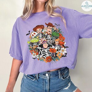 May include: A light purple short-sleeve t-shirt featuring a graphic print of Toy Story characters dressed in skeleton costumes. The design includes Woody, Buzz Lightyear, Jessie, Rex, and an alien surrounded by orange pumpkins, flowers, and spiderwebs.
