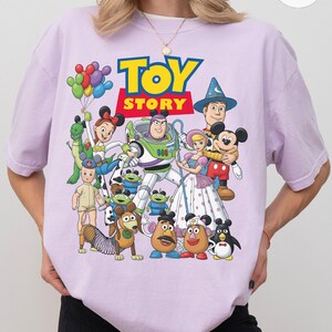 Op de afbeelding: Een lichtpaars T-shirt met de woorden "TOY STORY" in geel en blauw, met een kleurrijke afbeelding van personages uit de animatiefilm. Het ontwerp bevat Buzz Lightyear, Woody en Mickey Mouse.