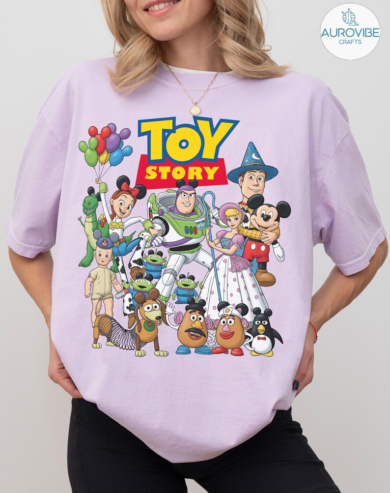Op de afbeelding: Een lichtpaars T-shirt met de woorden "TOY STORY" in geel en blauw, met een kleurrijke afbeelding van personages uit de animatiefilm. Het ontwerp bevat Buzz Lightyear, Woody en Mickey Mouse.