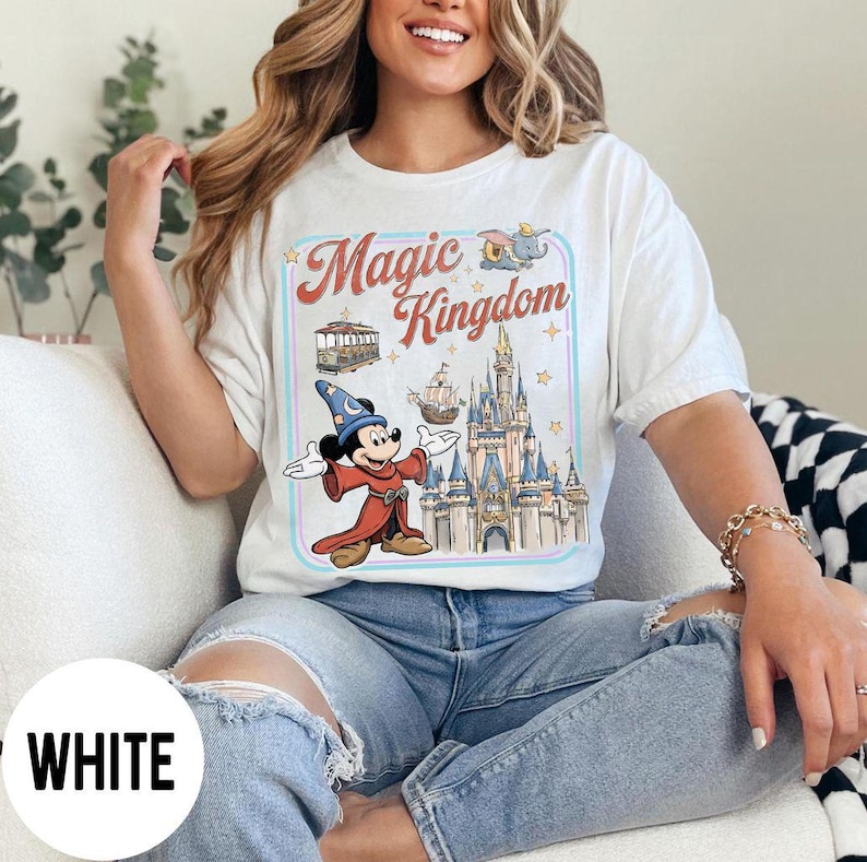 May include: White t-shirt featuring a graphic of Mickey Mouse in a wizard outfit, the Magic Kingdom logo, and a castle. The design includes a trolley, a ship, and stars. The shirt is a relaxed fit.
