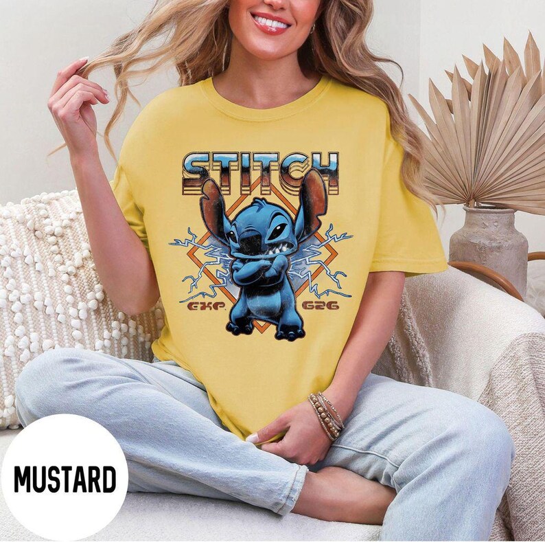 Puede incluir: Camiseta amarilla mostaza con un personaje de dibujos animados azul, orejas grandes y la palabra "STITCH" en una fuente retro. El personaje est&aacute; rodeado de rayos y el texto "GXP G26".