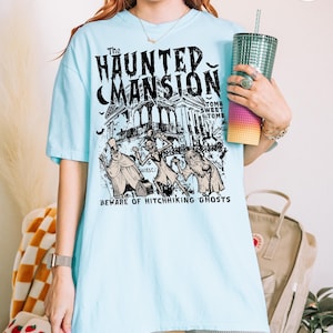 May include: Light blue t-shirt featuring a graphic of "The Haunted Mansion" with ghosts and the text "Beware of Hitchhiking Ghosts." The design is in black and white. The shirt is a short-sleeved, casual style.