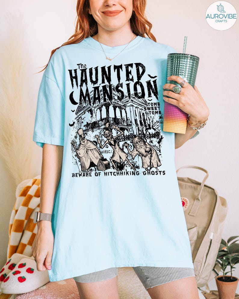 May include: Light blue t-shirt featuring a graphic of "The Haunted Mansion" with ghosts and the text "Beware of Hitchhiking Ghosts." The design is in black and white. The shirt is a short-sleeved, casual style.
