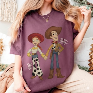 Może przedstawiać: Bordowa koszulka z kresk&oacute;wkową ilustracją Jessie i Woody'ego z Toy Story, trzymających się za ręce. Jessie ma na sobie białą koszulę, spodnie w krowie łaty i czerwony kapelusz. Woody ma na sobie kapelusz kowbojski, ż&oacute;łtą koszulę i niebieskie dżinsy. Na koszulce widnieją imiona "Jessie" i "Woody".