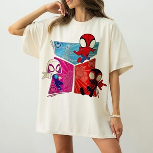 May include: Cream-colored t-shirt featuring a colorful graphic of Spider-Man characters. The design includes Spider-Man, Spider-Gwen, and Miles Morales in a geometric arrangement. The shirt has short sleeves and a relaxed fit.