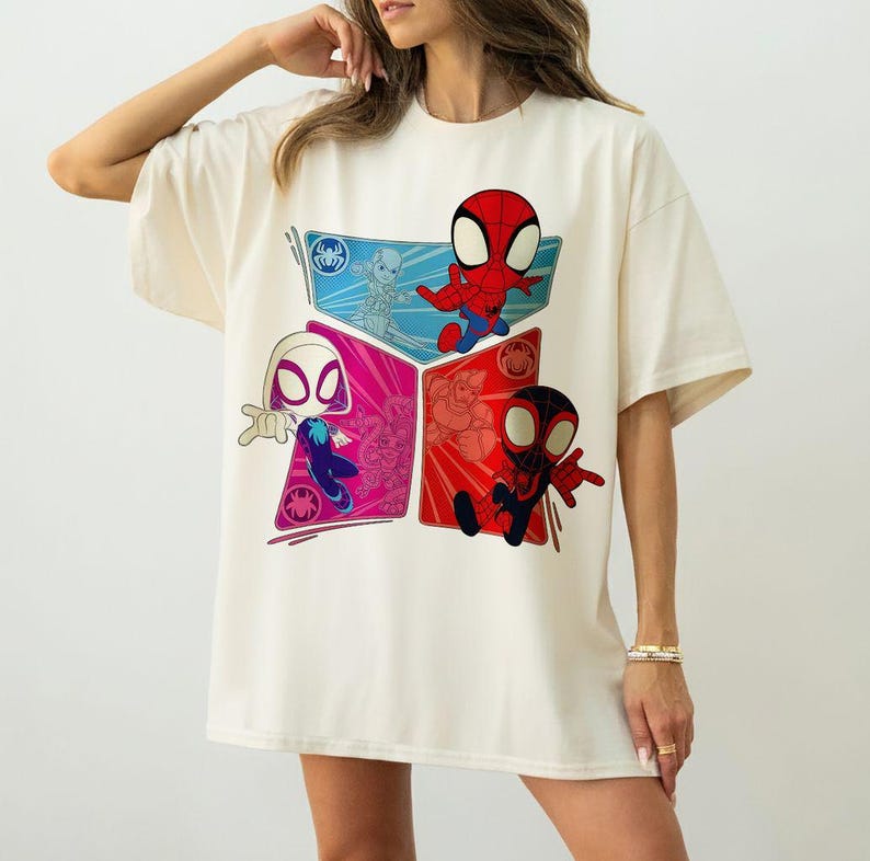 May include: Cream-colored t-shirt featuring a colorful graphic of Spider-Man characters. The design includes Spider-Man, Spider-Gwen, and Miles Morales in a geometric arrangement. The shirt has short sleeves and a relaxed fit.