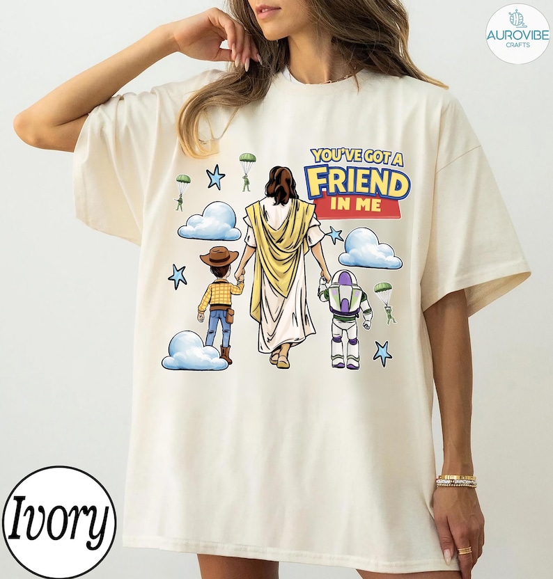 May include: Ivory-colored t-shirt featuring a graphic with the text "YOU'VE GOT A FRIEND IN ME." The design includes illustrations of Jesus, Woody, and Buzz Lightyear, with clouds, stars, and parachutes. The shirt has a relaxed fit.