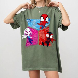 May include: Olive green t-shirt featuring a colorful graphic of Spider-Man characters. The design includes four sections with Spider-Man, Spider-Gwen, and other characters in red, blue, and pink. The shirt has short sleeves and a crew neck.