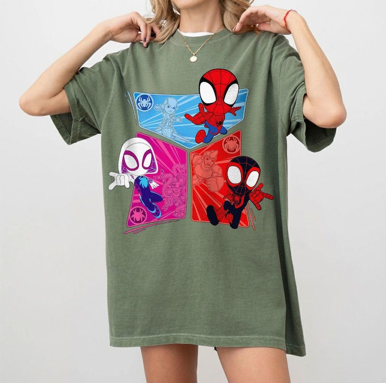 May include: Olive green t-shirt featuring a colorful graphic of Spider-Man characters. The design includes four sections with Spider-Man, Spider-Gwen, and other characters in red, blue, and pink. The shirt has short sleeves and a crew neck.