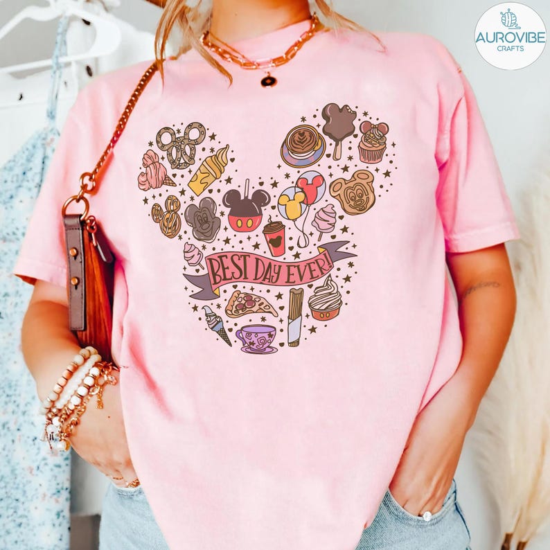 May include: A light pink t-shirt featuring a graphic of various snacks and treats arranged in the shape of a mouse head silhouette. A banner across the center contains the text "BEST DAY EVER" in a dark font, surrounded by small stars and dots.