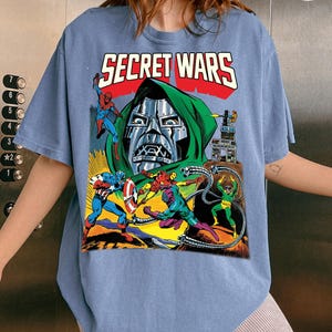 May include: A light blue t-shirt featuring a comic book graphic with the text "SECRET WARS" in red and white. The graphic depicts several comic book characters, including Doctor Doom, Spider-Man, and Captain America, in a colorful action scene.