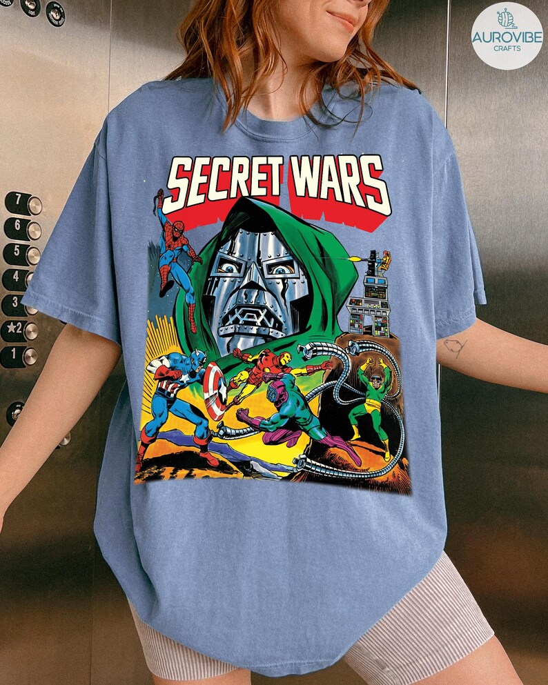May include: A light blue t-shirt featuring a comic book graphic with the text "SECRET WARS" in red and white. The graphic depicts several comic book characters, including Doctor Doom, Spider-Man, and Captain America, in a colorful action scene.