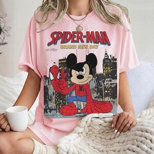 May include: A light pink t-shirt featuring a graphic of Mickey Mouse dressed as Spider-Man, with the words "SPIDER-MAN BRAND NEW DAY" in red and blue. The design includes a cityscape background.