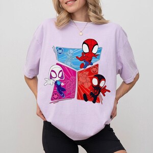 May include: A light purple t-shirt featuring a colorful graphic of three Spider-Man characters in a cartoon style. The characters are in red, black, and white, each within a different colored geometric shape. The shirt has short sleeves and a crew neck.