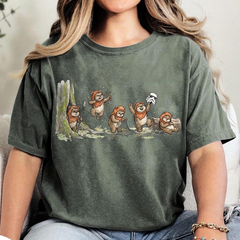 May include: Olive green t-shirt featuring a whimsical illustration of Ewoks in various poses. The cartoon Ewoks are depicted with spears, slingshots, and a net, and one is holding a Stormtrooper helmet. The design is set against a forest backdrop.