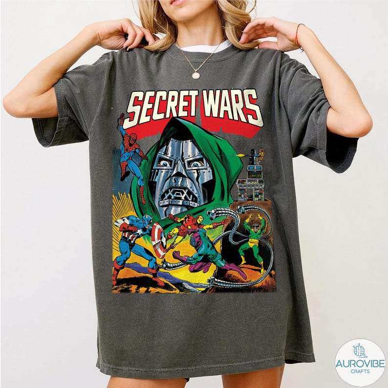 May include: Dark gray t-shirt featuring a comic book illustration with the text "SECRET WARS". The graphic includes characters like Spider-Man, Captain America, and Doctor Doom in a colorful action scene.
