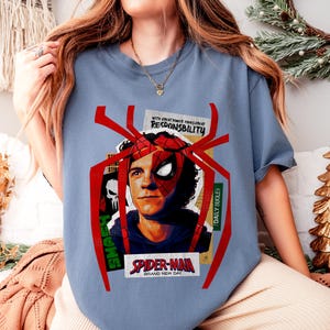 Op de afbeelding: Lichtblauw T-shirt met een Spider-Man-afbeelding. Het ontwerp bevat een portret van het personage en de tekst "SPIDER-MAN BRAND NEW DAY". De tekst "WITH GREAT POWER COMES GREAT RESPONSIBILITY" is ook zichtbaar.