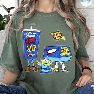 以下が含まれることがあります： ピザプラネットのデザインが施されたグリーンのTシャツ。デザインには、青いメガガルのプカップ、ピザプラネット、スーパーノヴァバーガーの箱、緑色のエイリアンキャラクターが含まれています。Tシャツのテキストは「Pizza Planet」、「Mega Gulp」、「Super Nova Burger」と書かれています。