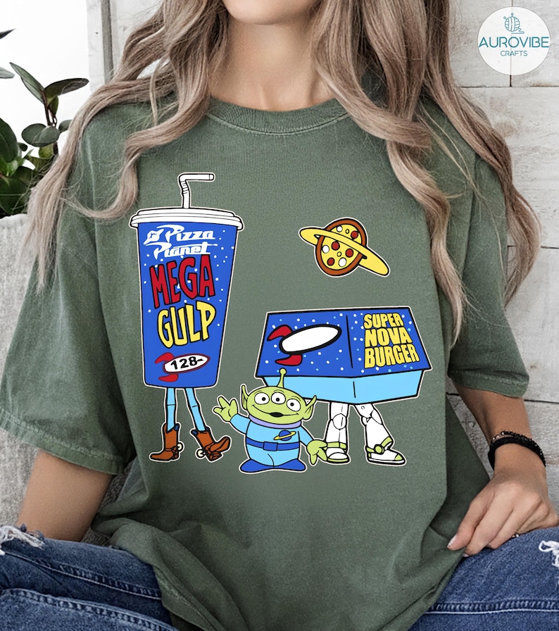 以下が含まれることがあります： ピザプラネットのデザインが施されたグリーンのTシャツ。デザインには、青いメガガルのプカップ、ピザプラネット、スーパーノヴァバーガーの箱、緑色のエイリアンキャラクターが含まれています。Tシャツのテキストは「Pizza Planet」、「Mega Gulp」、「Super Nova Burger」と書かれています。