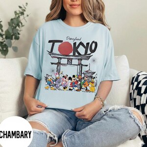 Puede incluir: Camiseta azul claro con la inscripci&oacute;n "Disneyland Tokyo", un c&iacute;rculo rojo y un dise&ntilde;o de puerta japonesa. La camiseta presenta personajes de dibujos animados como Mickey Mouse, Donald Duck y Goofy.