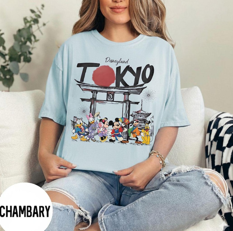 Puede incluir: Camiseta azul claro con la inscripci&oacute;n "Disneyland Tokyo", un c&iacute;rculo rojo y un dise&ntilde;o de puerta japonesa. La camiseta presenta personajes de dibujos animados como Mickey Mouse, Donald Duck y Goofy.