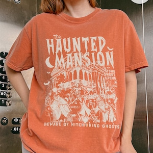 May include: Orange t-shirt featuring a graphic of The Haunted Mansion with the text "Beware of Hitchhiking Ghosts." The design includes a mansion, bats, and figures. The shirt is a crew neck.