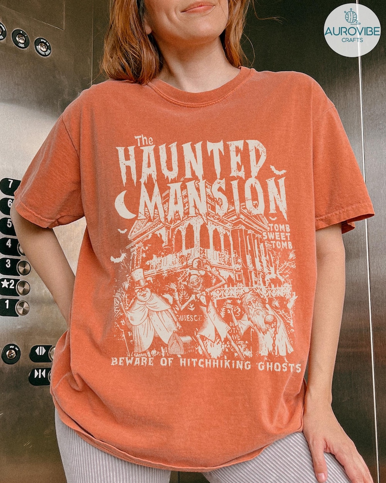 May include: Orange t-shirt featuring a graphic of The Haunted Mansion with the text "Beware of Hitchhiking Ghosts." The design includes a mansion, bats, and figures. The shirt is a crew neck.